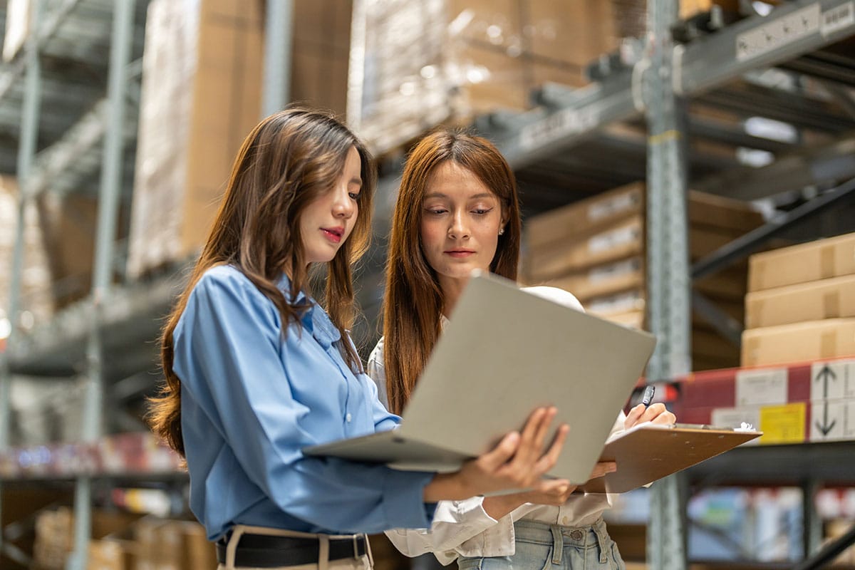 Two logistics professionals using a laptop to track inventory inside a warehouse with high shelves and cardboard boxes.