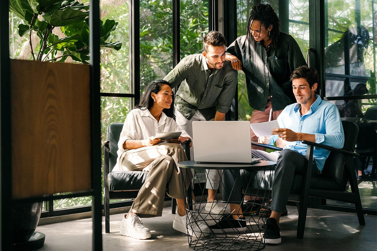A diverse team of professionals collaborating around a laptop and documents in a bright, modern workspace filled with plants.