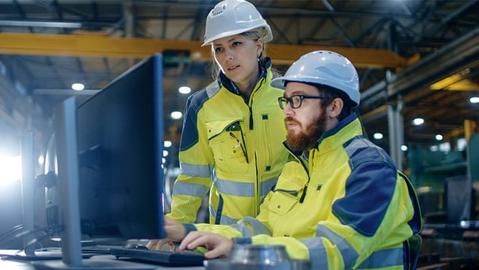 Two workers in a factory, wearing hard hats and yellow safety vests, collaborate while looking at a laptop.