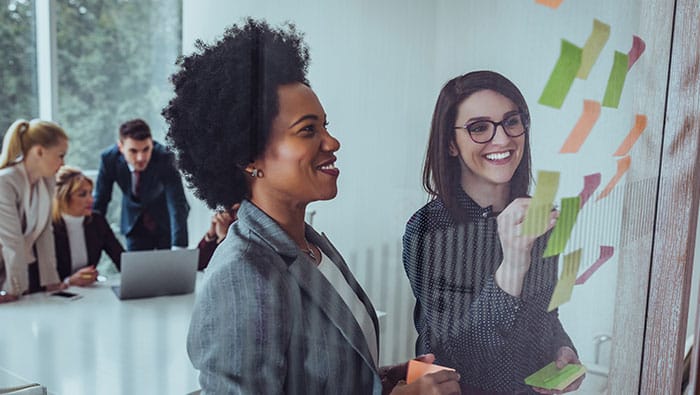 A diverse team collaborates in a workshop, using sticky notes on a glass wall to design a secure IoT system and strategize network requirements.